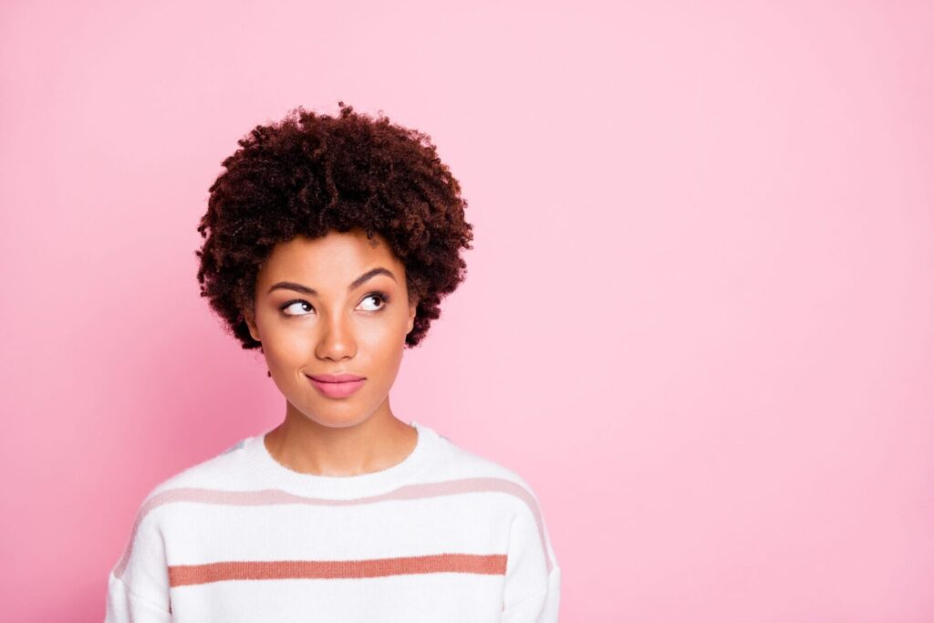 Photo of curious sly black skinned girl glancing at empty space wearing striped white sweater planning her life isolated over pink pastel color background